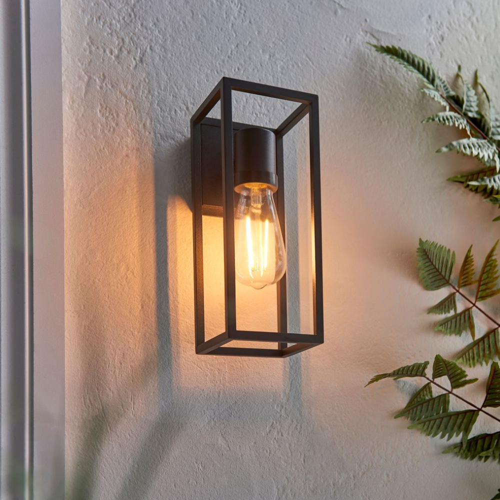 Wall sconce with exposed bulb in black metal frame, casting warm glow on textured white wall with fern leaves.