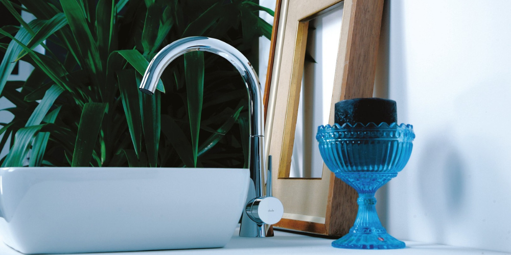 Bathroom sink with chrome swivel spout tap, plant, black candle in blue glass holder, and empty wooden picture frame.