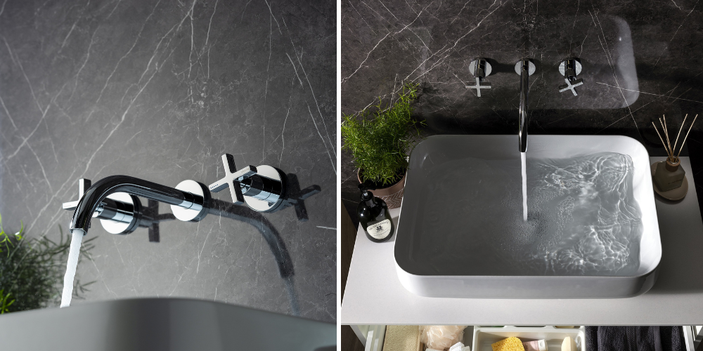 Wall-mounted basin mixer taps on dark marble wall, water flowing into white rectangular sink with plants and toiletries.
